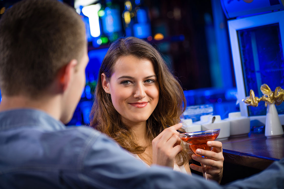 Frau mit Cocktail in der Hand lächelt Mann an der Bar an